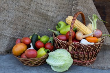 Olgun sebzeler, salatalıklar, biberler, soğanlar, sarımsak, mısır, pancar, patlıcan, domatesler sepette ve hasır bir tabakta yatıyor. Yakınlarda bir lahana başı yatıyor.