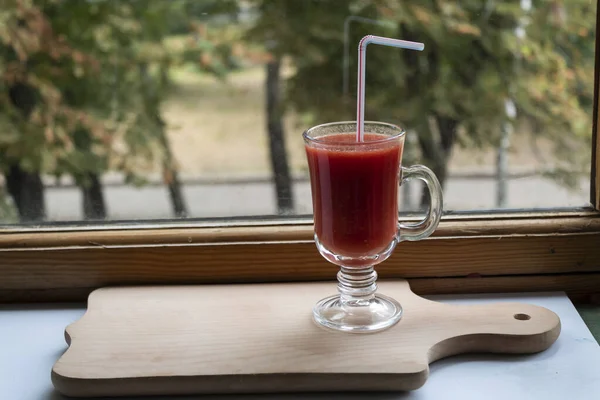 On the windowsill, on a cutting board, in a glass, there is red tomato juice with a straw.