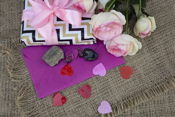 On the table are pink roses, a gift box, an envelope, earrings and two natural semi-precious stones, black onyx and arsenopyrite
