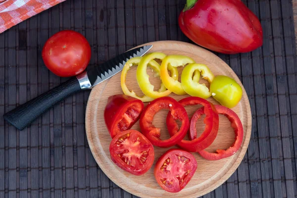 There are two types of chopped peppers and tomato slices on the round board. Nearby lies a knife and a whole pepper and tomato