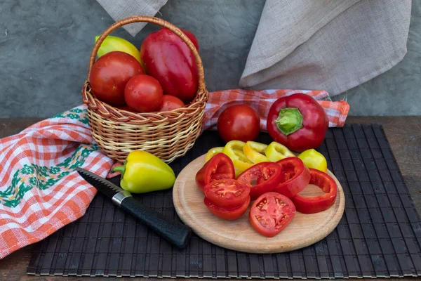 There are two types of chopped peppers and tomato slices on the round board. Nearby lies a knife, whole peppers and tomatoes, and a basket of vegetables