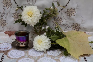 Bardak tutacağında beyaz bir mendilin üzerinde bir bardak çay var. Bir vazonun içinde bir buket beyaz yıldız çiçeği ve kar çileği var. Tepside sarı bir yaprak ve çiçekler var..