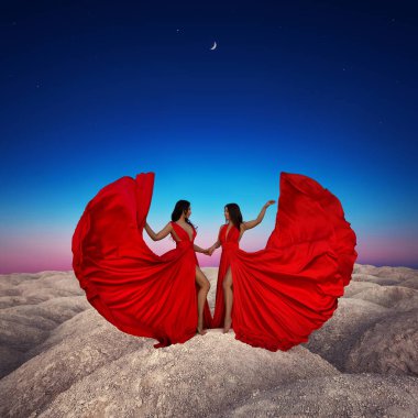 Birbirinin aynı kırmızı ipek elbiseli iki güzel esmer kız geceleri kum tepelerinin arka planında..