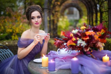 Leylak rengi elbiseli güzel bir kızın portresi sonbahar bahçesinde masa örtüsü, parlak kırmızı bir buket ve mumlarla çay içiyor. Romantik akşam fotoğrafı..