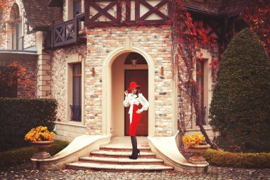 Kırmızı şapkalı, beyaz bluzlu ve kırmızı etekli güzel bir genç kız sonbahar bahçesinde güzel bir sanat kulübesinin kapısında duruyor..