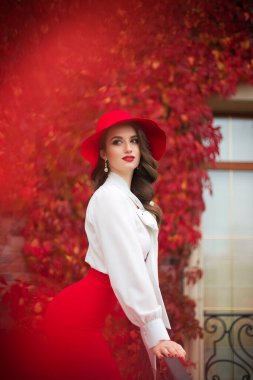Kırmızı şapkalı, beyaz bluzlu ve kırmızı etekli güzel bir kız kırmızı yaprakların arkasında duruyor.