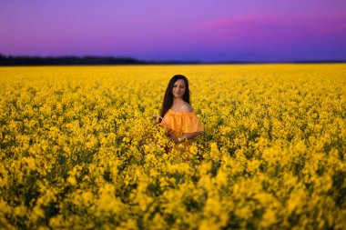 Sarı uzun elbiseli güzel bir esmer kız akşam gökyüzüne karşı sarı kolza tohumu tarlasında duruyor.