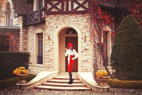 Kırmızı şapkalı, beyaz bluzlu ve kırmızı etekli güzel bir genç kız sonbahar bahçesinde güzel bir sanat kulübesinin kapısında duruyor..