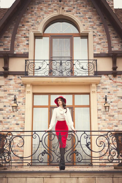 A beautiful young girl in a red hat, white blouse and red skirt standing on the terrace of the cottage