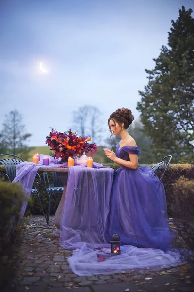 Beautiful young girl in a lilac dress drinking tea in an autumn garden at a table with a lilac tablecloth and a red bouquet, in the light of a Crescent moon and candles. Romantic evening photo.