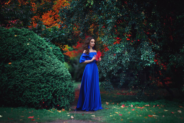 Romantic portrait of a beautiful young girl with blue makeup and red lips, in a long ultramarine blue dress with long blue earrings, on the background of an autumn ark with red leaves