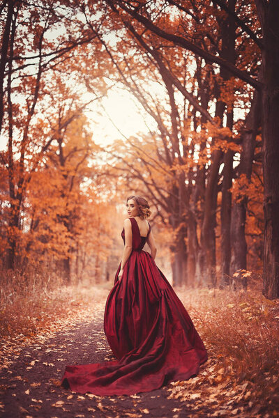 Romantic portrait of a young girl in a long red dress standing in an autumn alley
