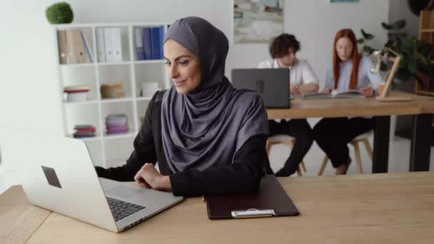 Mulher muçulmana bebendo café e tendo uma conversa em vídeo em um laptop no escritório — Vídeo de Stock