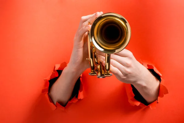Mãos tocando trompete através de buracos rasgados no fundo papel vermelho — Fotografia de Stock