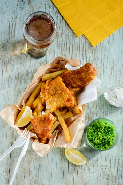 Deep fried fish and chips in combination with lemon slices, green peas and beer
