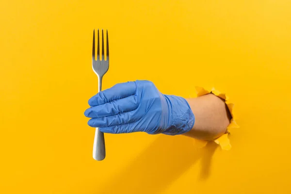 Mână într-o mănușă din latex care ține o furculiță metalică prin perete galben — Fotografie, imagine de stoc