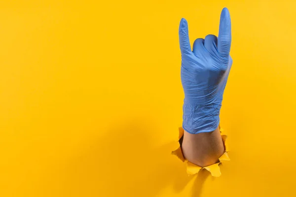 Hand in a latex glove showing a sign of the horns through torn yellow wall — Stock Photo, Image
