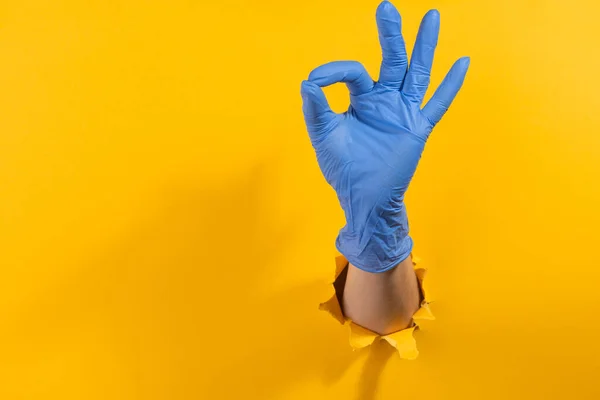 Hand in a latex glove showing an alright sign through torn yellow background — Stock Photo, Image