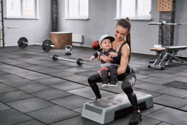 Küçük çocuğuyla spor salonunda çömelme hareketi yapan bir kadın.