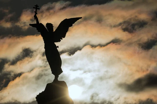 Silhouette of an angel with a cross in his hand on a background of dramatic sky and sun. Fragment of the Monument Mermaid , installed in 1902 in the Kadriorg park