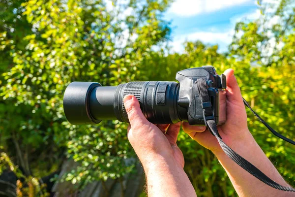 Digital SLR camera in the hands of a man on a background of greenery on a clear sunny day. Photo hunting in nature.