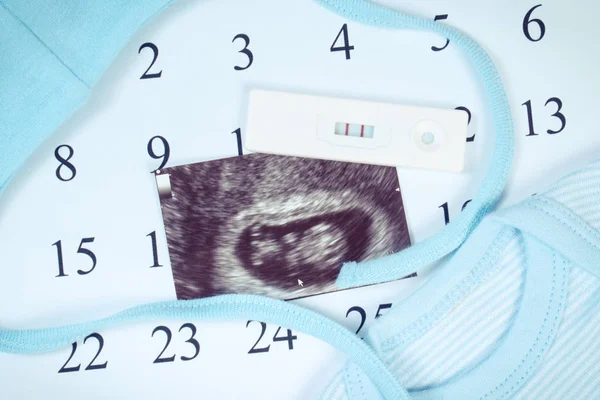 Pregnancy test with positive result, ultrasound scan of baby and clothing for newborn on calendar