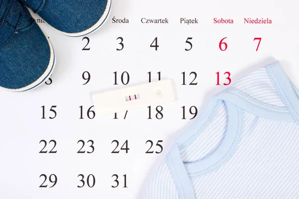 Тест на беременность с положительным результатом и одеждой для новорожденного в календаре, концепция ожидания ребенка — стоковое фото