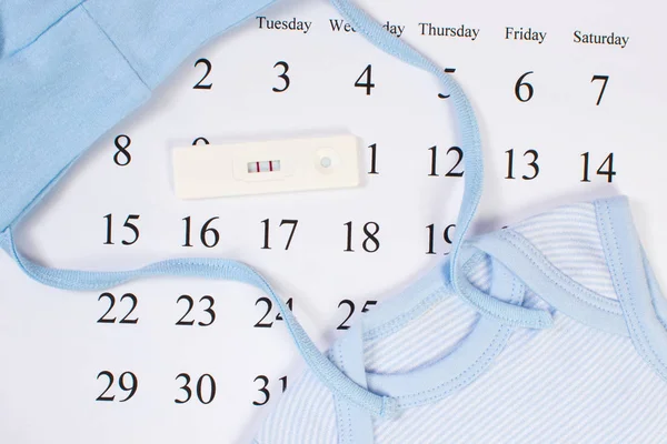 Test di gravidanza con risultato positivo e abbigliamento per neonato sul calendario, in attesa per il bambino — Foto Stock