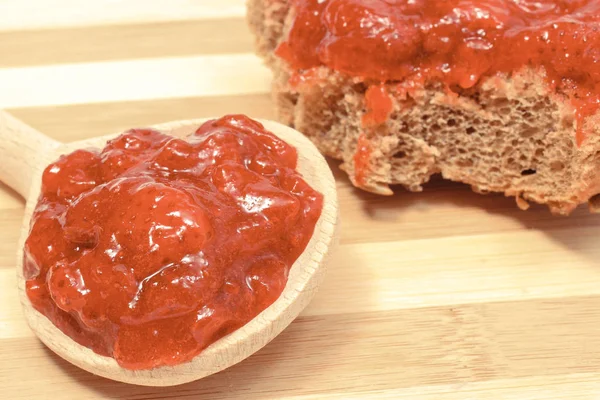 Strawberry jam on wooden spoon and wheat or rye bread in background — Stock Photo, Image
