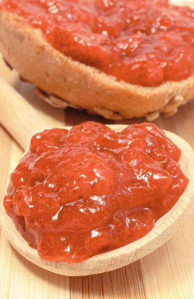 Strawberry jam on wooden spoon and whole wheat bread in background — Stock Photo, Image