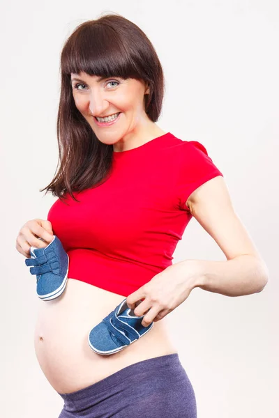 Donna sorridente incinta di scarpe per bambini, in attesa per il bambino — Foto Stock