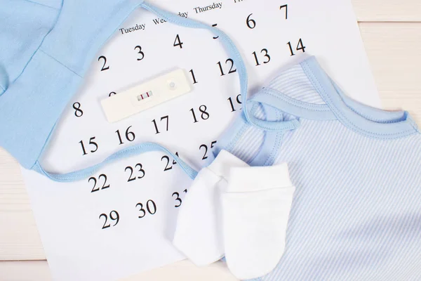 Test di gravidanza con risultato positivo e abbigliamento per il neonato sul calendario, in attesa del concetto di bambino — Foto Stock
