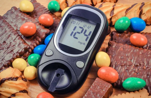 Glucose meter for checking sugar level and heap of candies with cookies. Diabetes and reduction of eating sweets