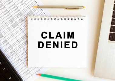 Bilgisayarın, hesap makinesinin ve ofis malzemelerinin yanındaki beyaz arka planda CLAIM DENIED yazılı not defteri. İş konsepti.