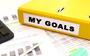 Sarı belge klasörü ofis masasının üzerinde. Hesap makinesi ve finans grafiklerinin yanında. İfade Hedeflerim.