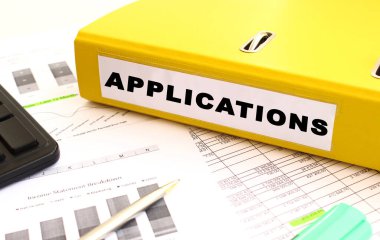 Sarı belge klasörü ofis masasının üzerinde. Hesap makinesi ve finans grafiklerinin yanında. Yazıt Uygulamaları.