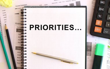 Hesap makinesi ve ofis malzemelerinin yanında beyaz bir arkaplan üzerinde PRIORITIES yazılı not. İş konsepti.
