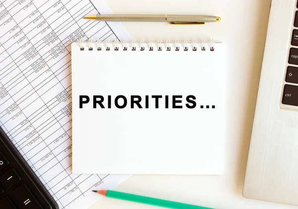 Bilgisayarın, hesap makinesinin ve ofis malzemelerinin yanında beyaz bir arkaplanda PRIORITIES mesajı olan bir not. İş konsepti.