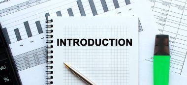 Ofis masasındaki mali tabloların üzerinde duran not defterinin sayfasına INTRODUCTION 'u yaz. Hesap makinesi ve işaretleyicinin yanında.