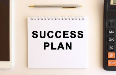 Hesap makinesi, tablet ve kalemin yanında beyaz bir arkaplan üzerinde SUCCESS PLAN metin ile not.