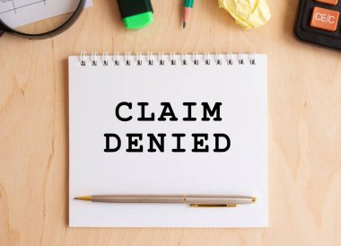Not defterinde CLAIM DENIED yazan ahşap bir masa, hesap makinesi ve ofis malzemeleri yanında.