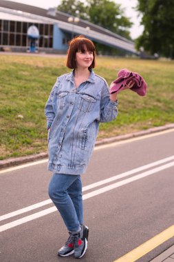 Kot pantolonlu bir kız kaldırımda duruyor, elinde pembe örgü bir eşarp tutuyor, keyfi yerinde ve yüzünde gülümseme var.