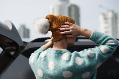 Kız kafasına kürk ponponu olan sarı örülü bir şapka takıyor, arka planda bir şehir manzarası..