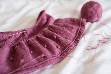 Yatağın üzerinde pembe örülmüş bir etek, beyaz bir çarşaftan beyaz bir arka plan, yün iplikler ve yakınlarda bir tüy yumağı var.