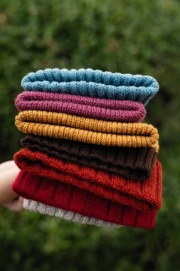 Örülmüş şapkalar yeşil çalıların arka planına karşı yığın halinde yatar. Elleriyle örgü şişleri ören renkli şapkalar. Bir yığın desenli kapaklar