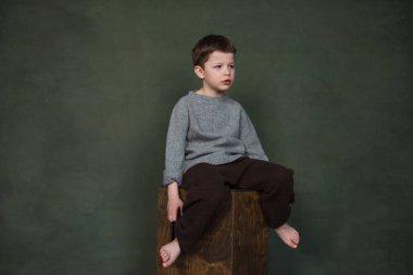 Örgü elbiseli yakışıklı bir çocuk ahşap bir kutunun üzerinde oturuyor.
