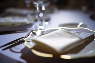 Akşam ışığı şenlik masasında, peçeteli güzel bir tabak ve restoranda bir menü, arka planda cam parlıyor.