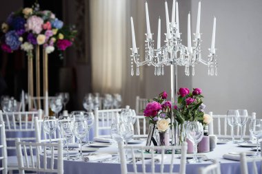 Restoranın beyaz salonunda düğün kutlaması için süslenmiş masalar var. Salonun süslemeleri taze çiçeklerle dolu. Masaların üzerinde uzun beyaz mumlar var.,