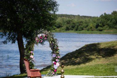 Taze çiçeklerle süslenmiş bir düğün kemeri nehir veya göle karşı bir parkta duruyor, yeşil çimenler, güneşli hava.,