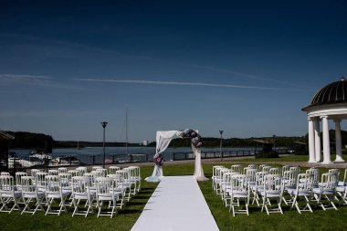 Nehir kenarındaki bir parkta düğün için dekorasyon, mavi gökyüzü, güneşli hava, beyaz kumaş ve taze çiçeklerle süslenmiş bir düğün kemeri, çimenlerin üzerinde konuk sandalyeler, yeni evliler için beyaz patika,